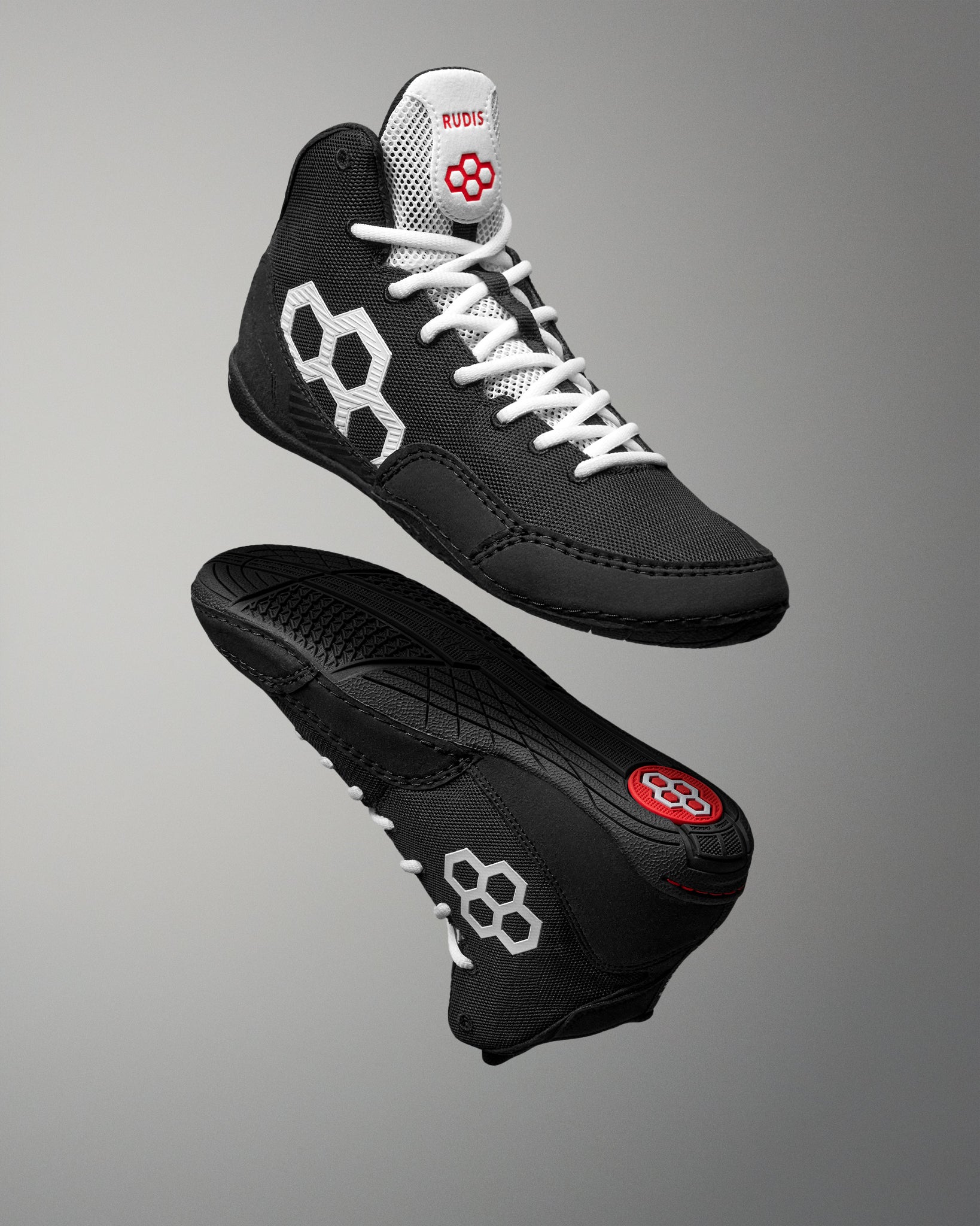 A pair of black athletic high-top wrestling shoes shown from the side and sole views against a gray gradient background. The shoes feature white laces, white geometric side logos, and a red logo on the tongue and heel.