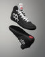 A pair of black athletic high-top wrestling shoes shown from the side and sole views against a gray gradient background. The shoes feature white laces, white geometric side logos, and a red logo on the tongue and heel.