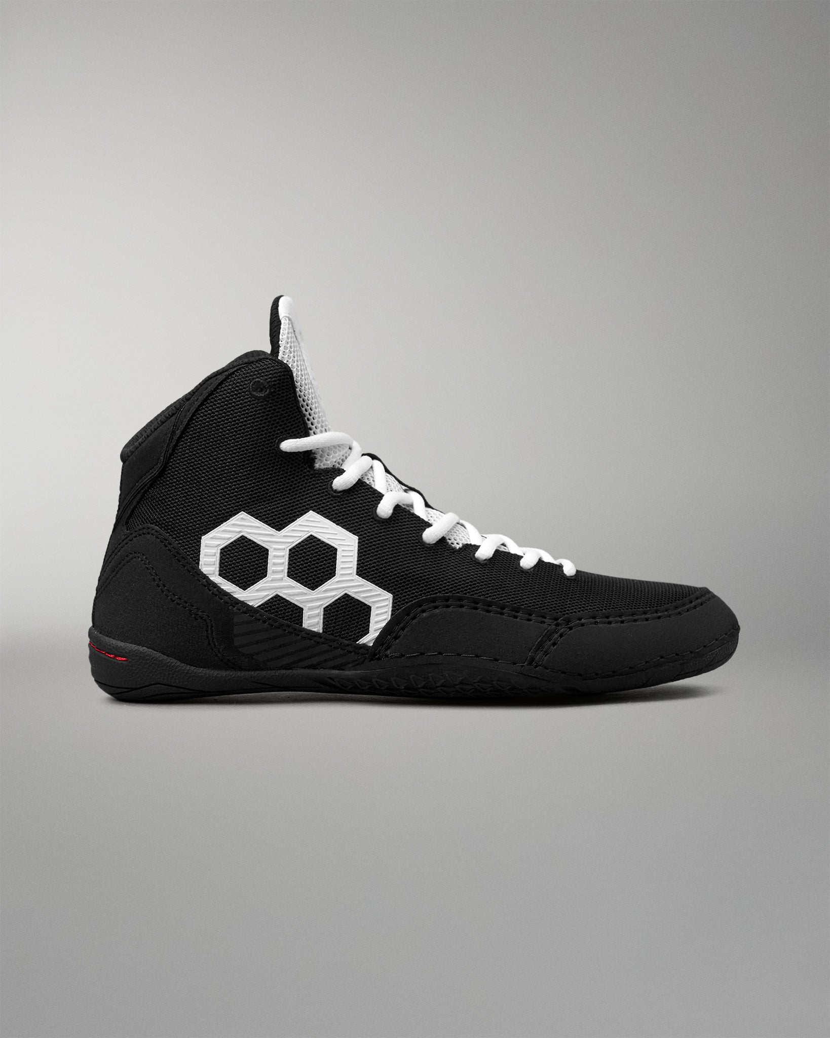 A black high-top wrestling shoe with white laces and a prominent white geometric hexagon logo on the side, shown against a neutral gray background.