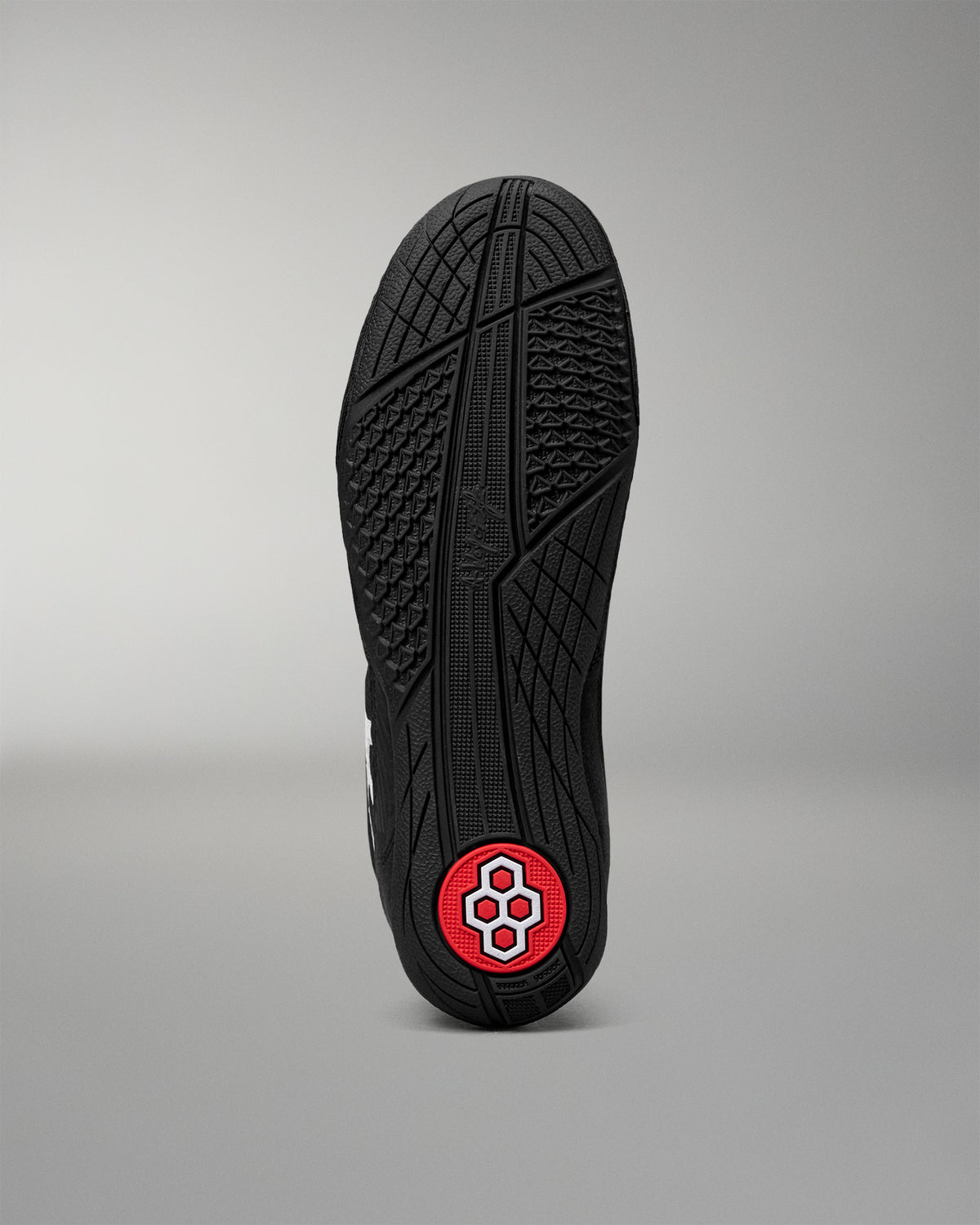 The black rubber outsole of a wrestling shoe, shown vertically against a neutral gray background. The sole features a textured tread pattern and a circular red heel patch with a white geometric logo.