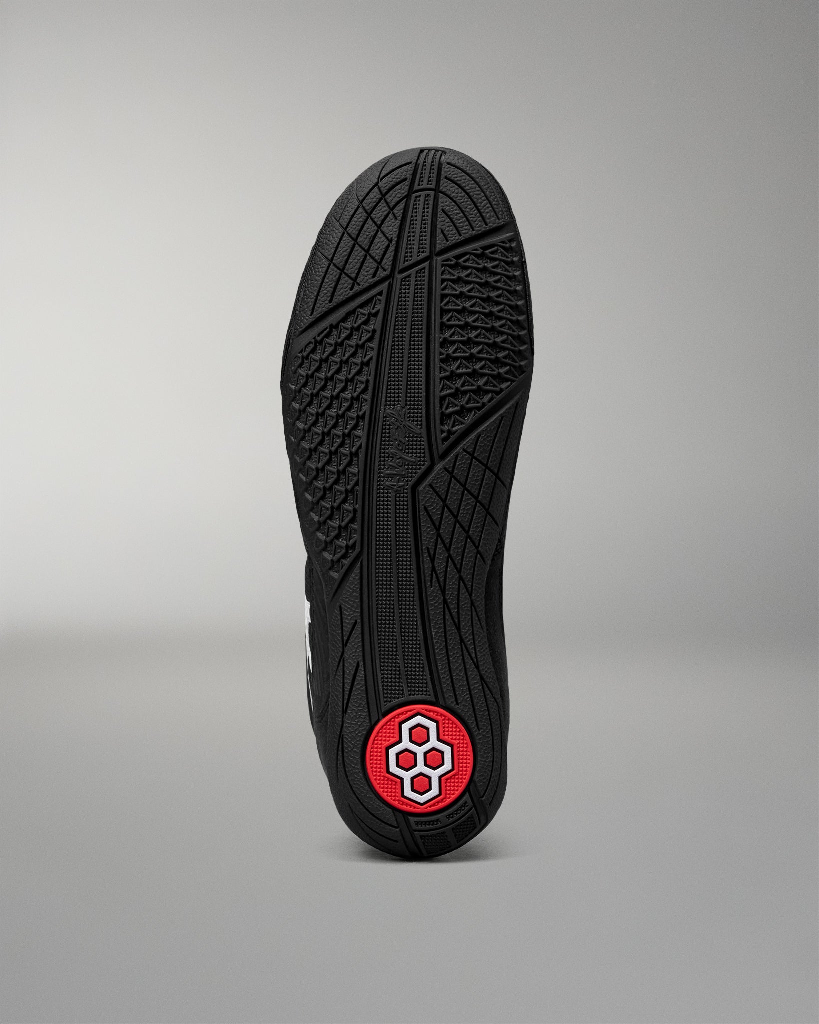 The black rubber outsole of a wrestling shoe, shown vertically against a neutral gray background. The sole features a textured tread pattern and a circular red heel patch with a white geometric logo.