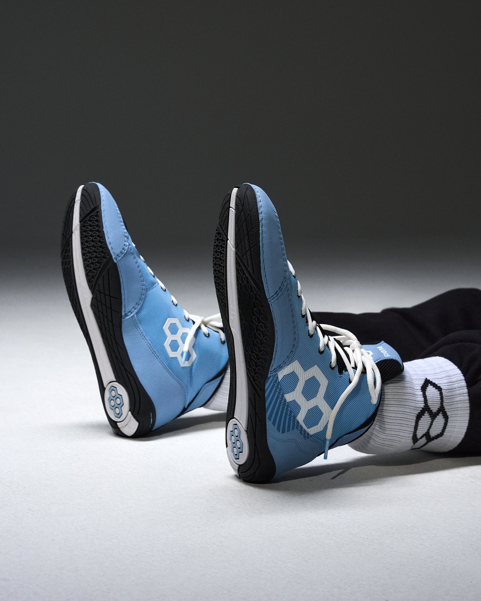 A pair of light blue and black high-top wrestling shoe with white geometric side logos, shown on a person's feet wearing white socks against a dark gray studio background.