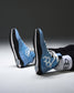 A pair of light blue and black high-top wrestling shoe with white geometric side logos, shown on a person's feet wearing white socks against a dark gray studio background.