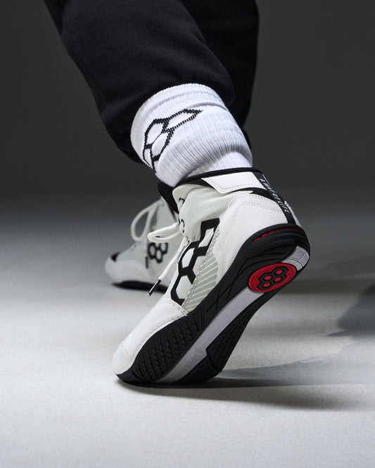 A close-up of a person's foot wearing a white, black, and red wrestling shoe, paired with white crew socks featuring a black logo, set against a dark gray studio background.