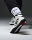 A close-up of a person's foot wearing a white, black, and red wrestling shoe, paired with white crew socks featuring a black logo, set against a dark gray studio background.