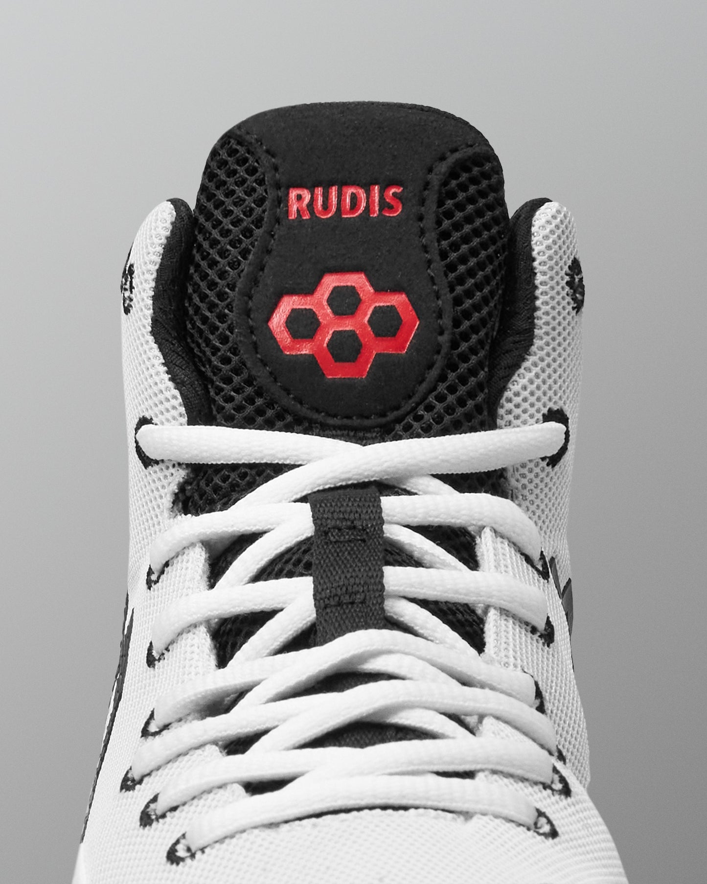 Close-up of a high-top wrestling shoe showing the tongue area, with a white mesh upper, black collar and tongue, white laces, and red embroidered branding that reads RUDIS with a red hexagon logo.
