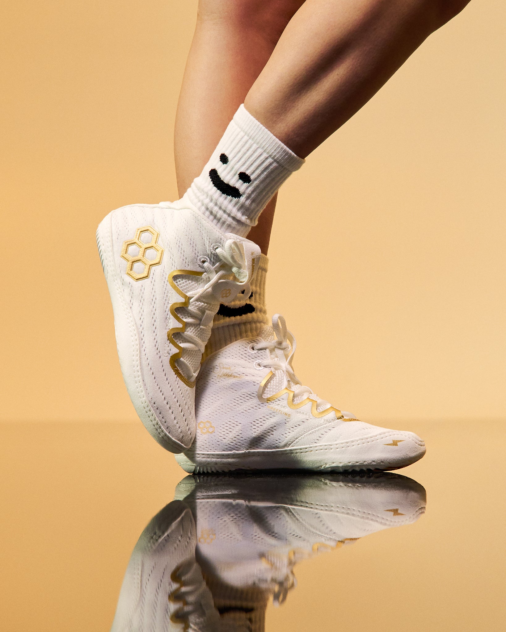 White athletic wrestling shoes with gold accents and white socks featuring a black smiley face, shown on a model's feet against a warm beige background with a reflective floor.