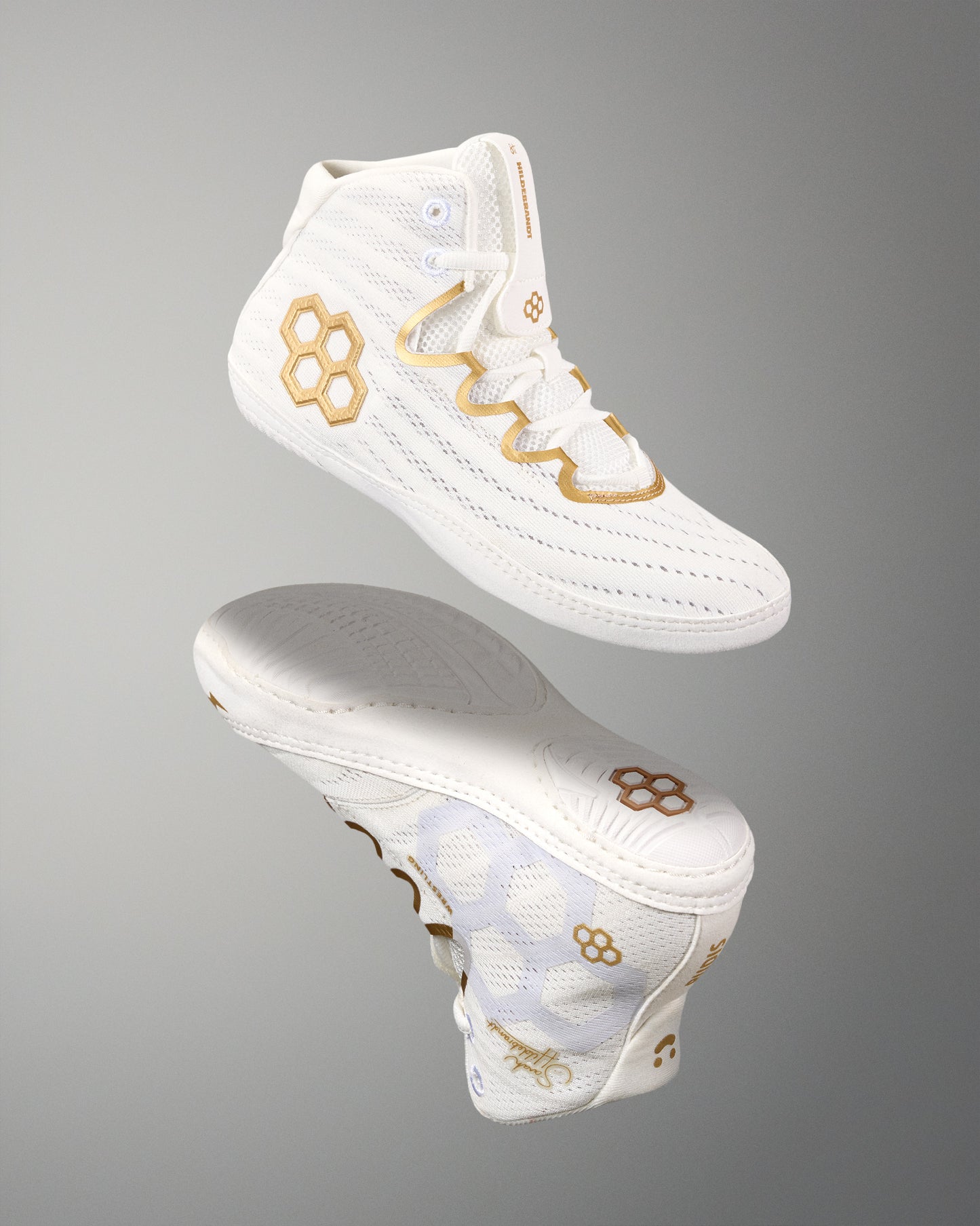 A pair of white wrestling shoes with gold quadhex and wavy geometric logo details, shown floating against a neutral gray gradient background.