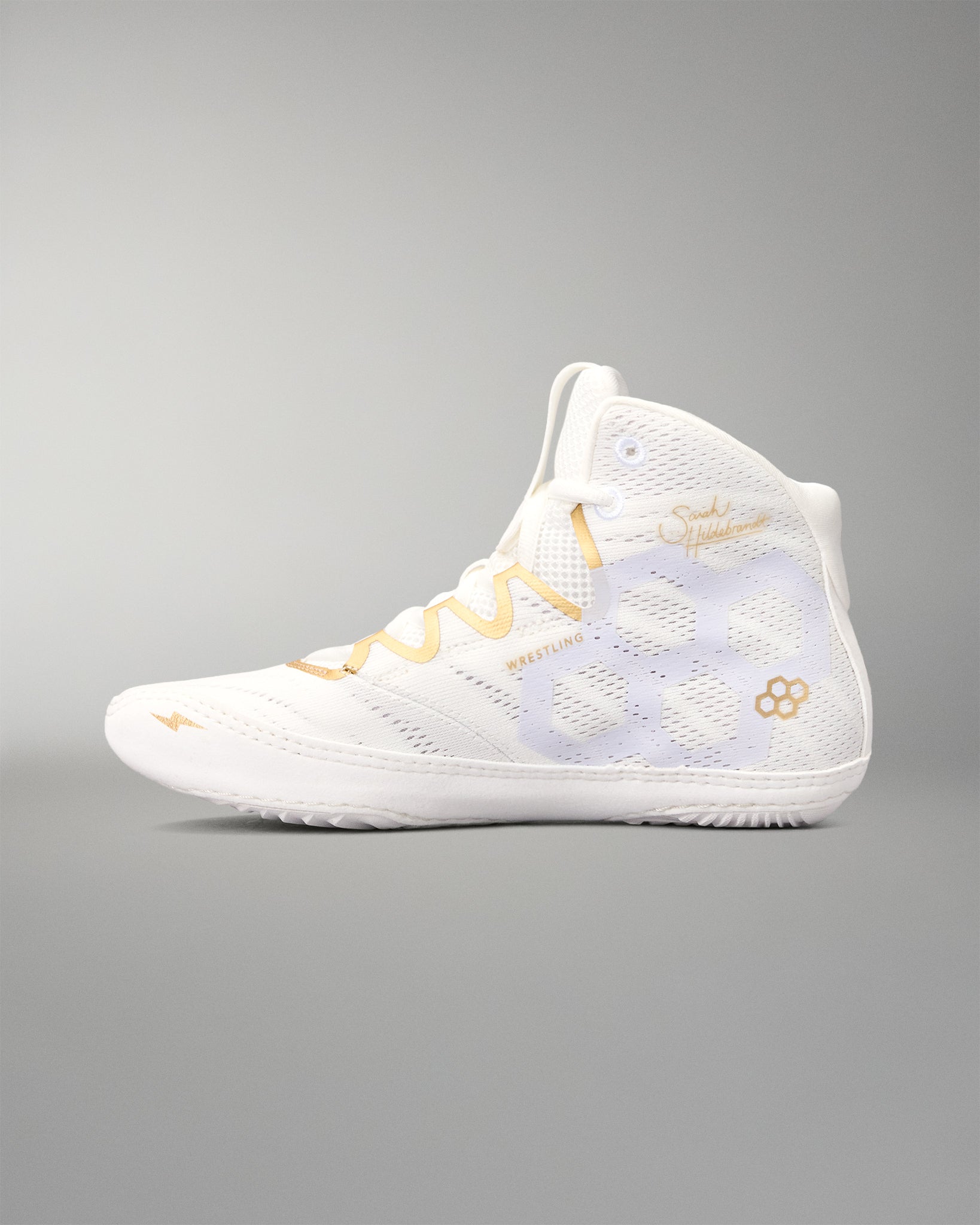 A high-top wrestling shoe in white with gold accents, including a wavy gold stripe on the laces, a gold flower logo on the side, and gold script text, shown against a neutral gray background.