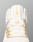 Close-up of the top of a white wrestling shoe with gold accents, featuring a white tongue strap with gold lettering, a gold geometric logo on the lace guard, and gold eyelets.