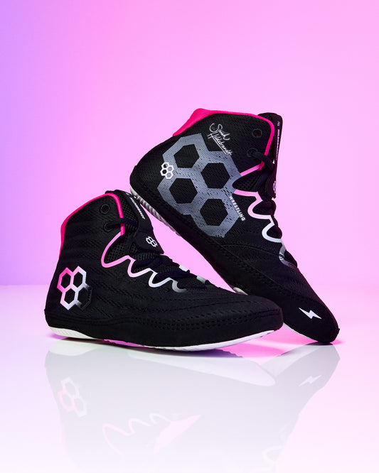 A pair of black high-top wrestling shoes with vibrant pink and gray accents, displayed on a reflective white surface against a pink gradient background.