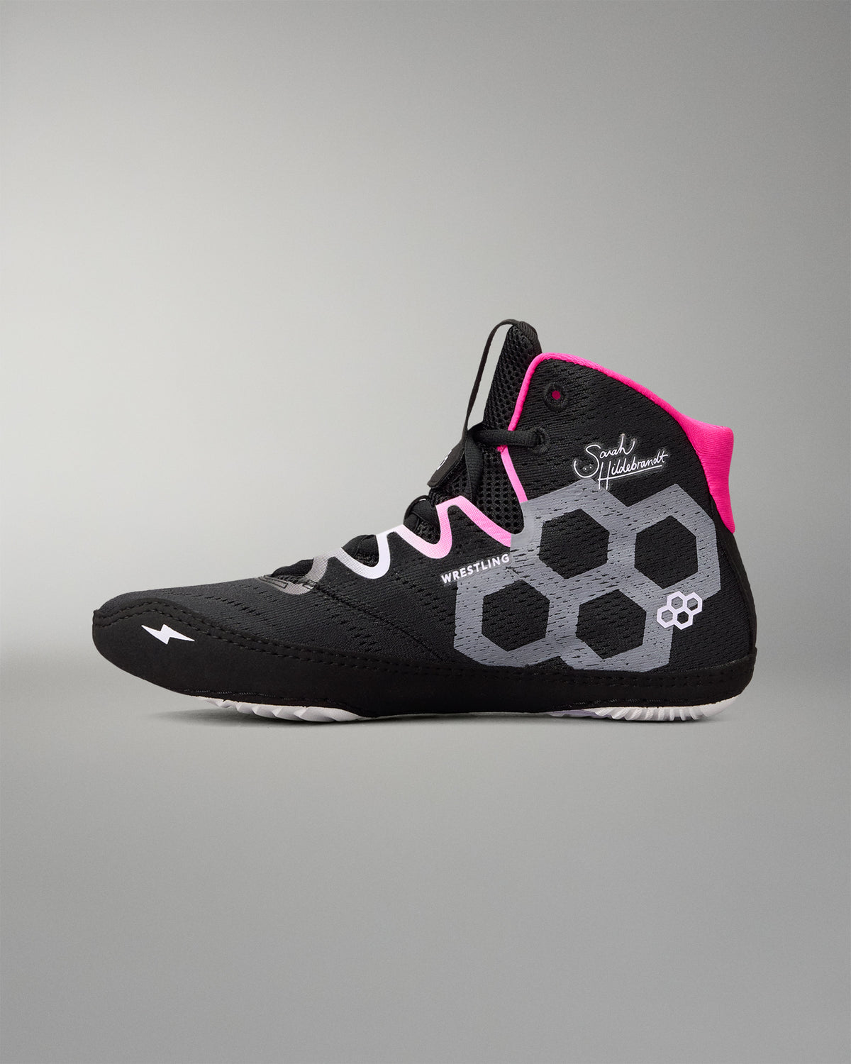 A high-top wrestling shoe in black with a gray honeycomb pattern, white accents, and bright pink details on the tongue and laces, shown against a neutral gray background.