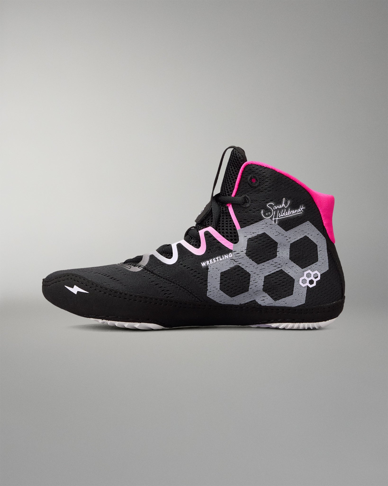 A high-top wrestling shoe in black with a gray honeycomb pattern, white accents, and bright pink details on the tongue and laces, shown against a neutral gray background.