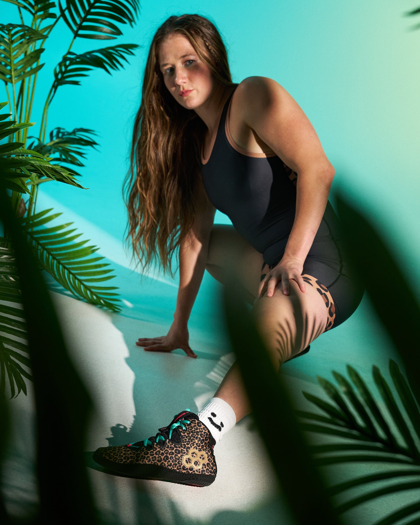 A woman in a black singlet and black shorts crouches in a tropical setting with palm leaves, wearing a leopard-print wrestling shoe and white socks.