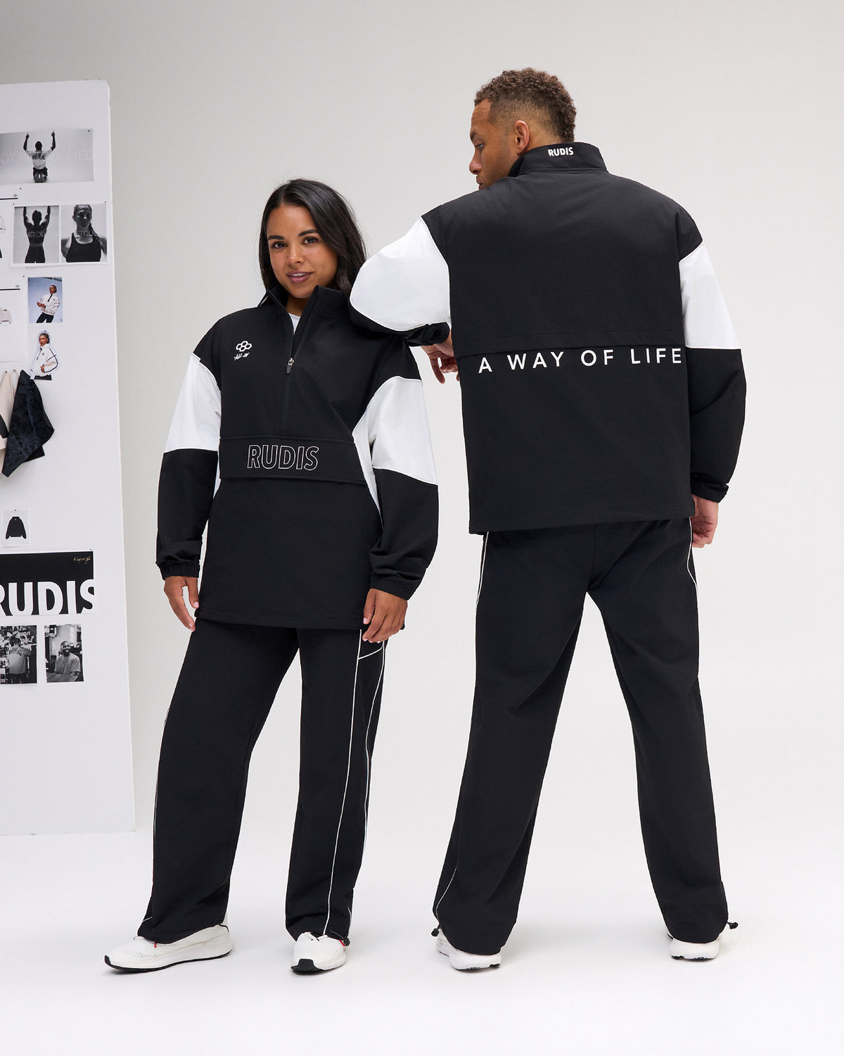 A man and a woman modeling matching black and white color-block tracksuits in a bright studio setting. The woman is shown from the front wearing a black half-zip jacket with white shoulder panels and 'RUDIS' printed on the chest, paired with black pants.