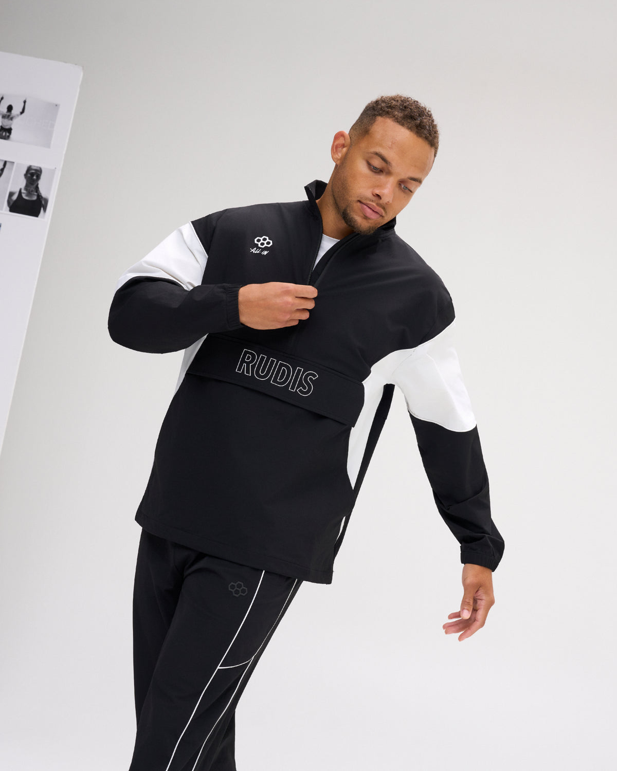 A man modeling a black and white color-blocked athletic jacket with 'RUDIS' printed on the chest, paired with matching black track pants with white piping, set against a clean light gray studio background.