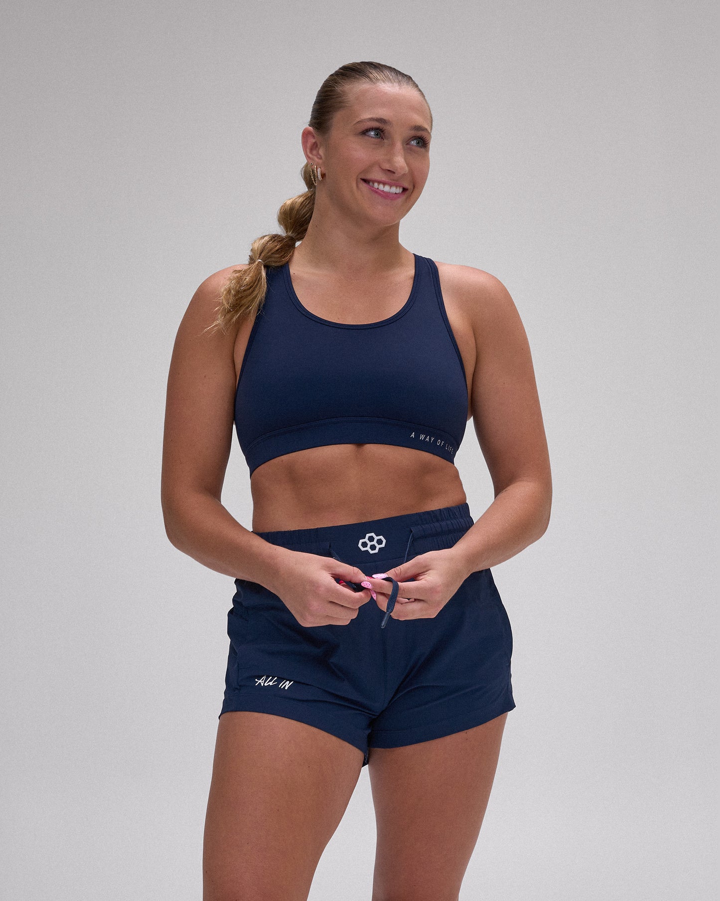A woman modeling a matching navy blue activewear set consisting of a sports bra and athletic shorts, photographed against a neutral gray studio background.