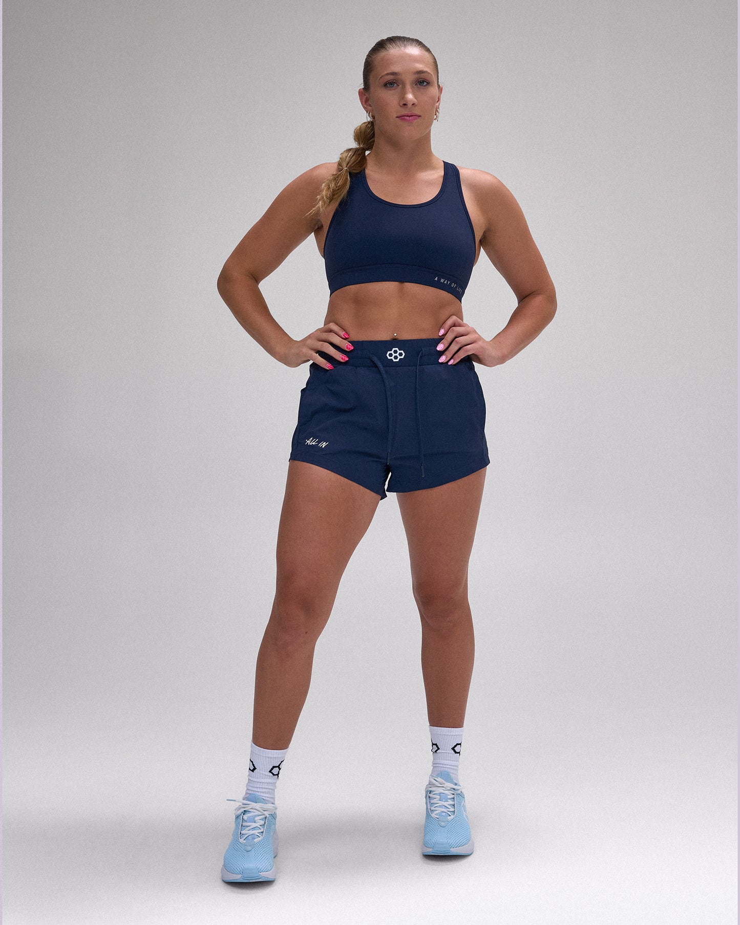 A woman modeling a navy blue activewear set consisting of a sports bra and matching shorts, paired with white socks and light blue sneakers, against a neutral gray studio background.