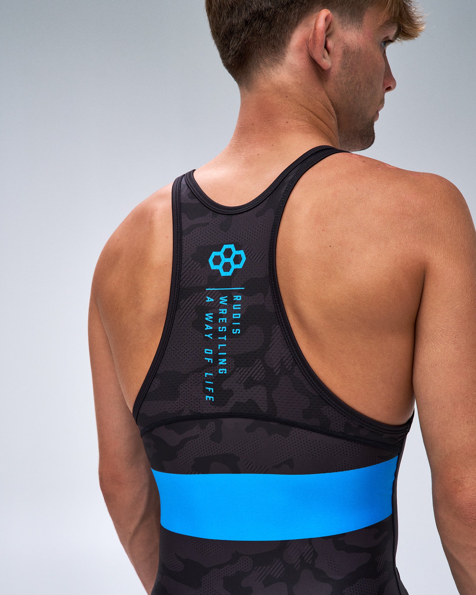 Rear view of a man wearing a black wrestling singlet with a bright blue horizontal band and blue printed logo and text on the back, against a light gray studio background.