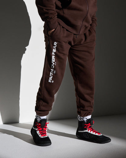 Person wearing a brown tracksuit with 'faith over fear' text and black wrestling shoes on a gray background