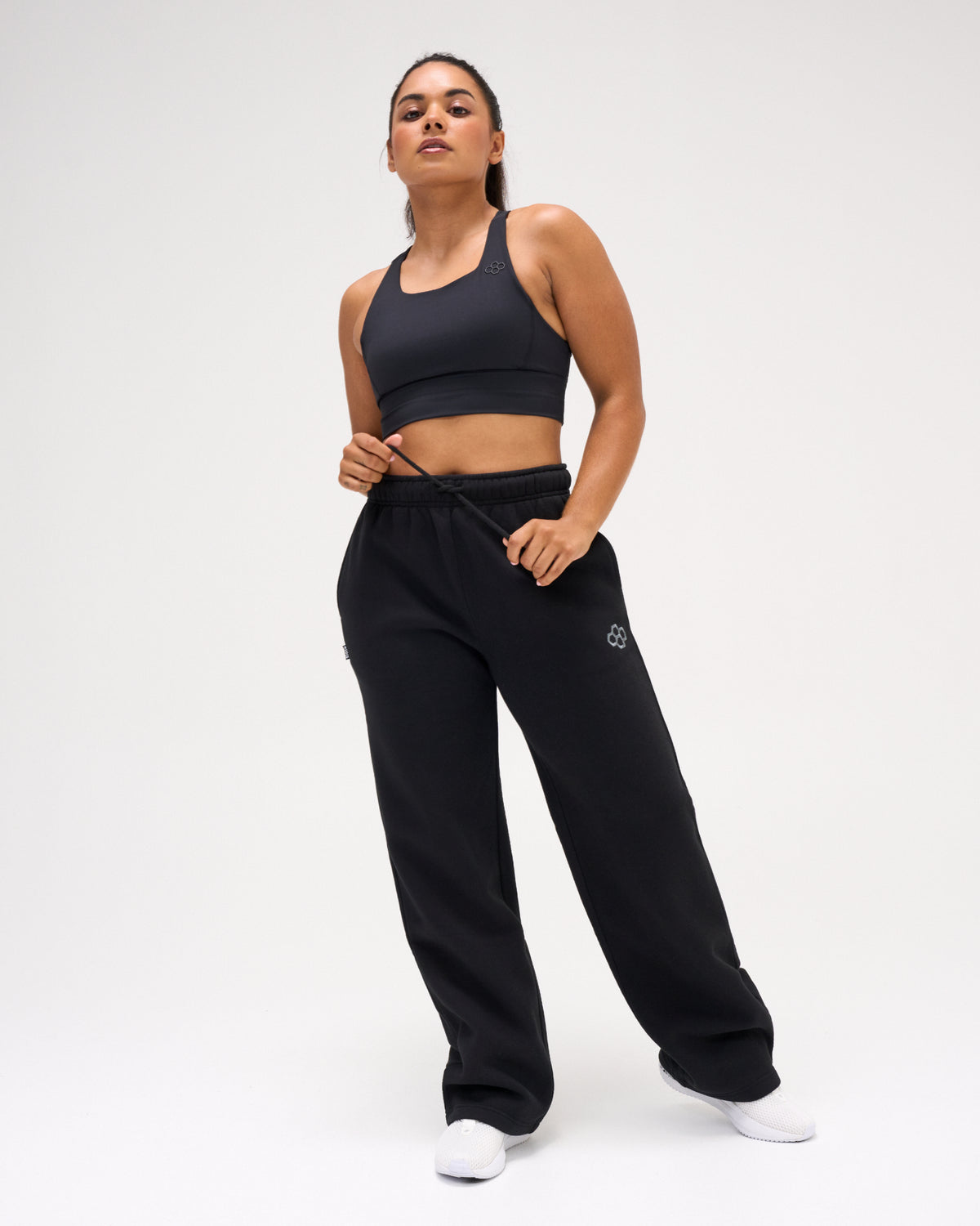 Female model wearing RUDIS Comfort Fleece Wide Leg Sweatpants in Black with a black athletic sports bra, showcasing the relaxed fit and adjustable drawstring waist. Copy Alt Text