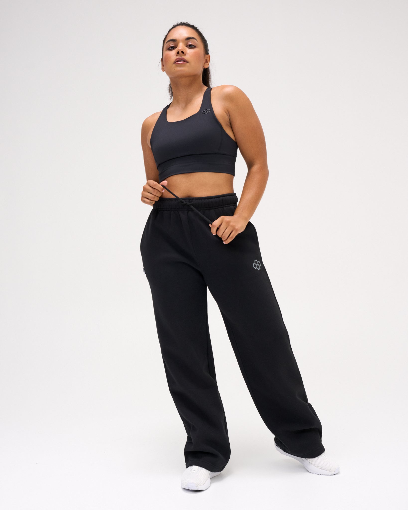 Female model wearing RUDIS Comfort Fleece Wide Leg Sweatpants in Black with a black athletic sports bra, showcasing the relaxed fit and adjustable drawstring waist. Copy Alt Text