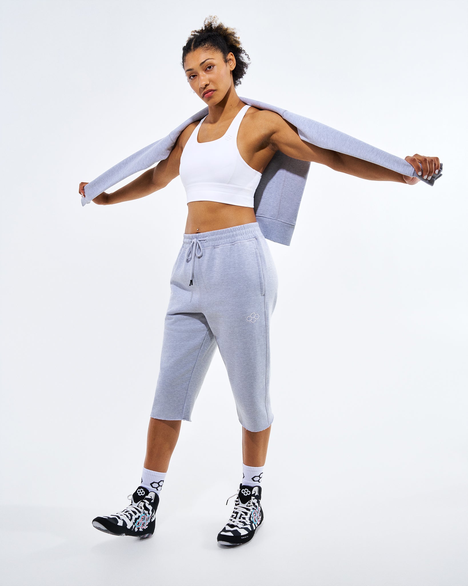 A woman modeling a light gray athletic set consisting of cropped sweatpants and a matching zip-up hoodie, worn over a white sports bra, with black and white sneakers, against a plain white studio background.