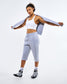 A woman modeling a light gray athletic set consisting of cropped sweatpants and a matching zip-up hoodie, worn over a white sports bra, with black and white sneakers, against a plain white studio background.