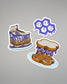 Three cartoon-style stickers of a cake, a skull, and a sandwich with purple sauce on a gray background.