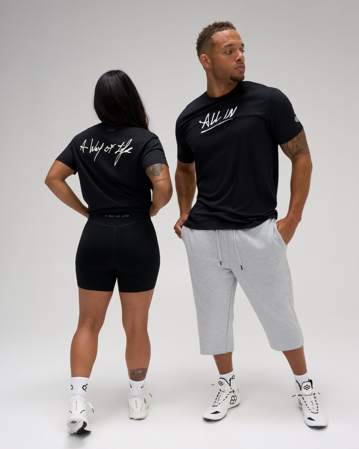 A man and a woman modeling black athletic apparel against a light gray studio background. The woman wears black biker shorts and a black t-shirt with white script text on the back.