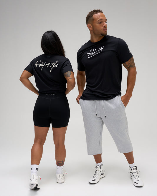 A man and a woman modeling black athletic apparel against a light gray studio background. The woman wears black biker shorts and a black t-shirt with white script text on the back.