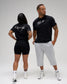 A man and a woman modeling black athletic apparel against a light gray studio background. The woman wears black biker shorts and a black t-shirt with white script text on the back.