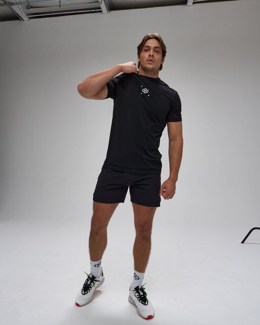 Man wearing a black t-shirt and shorts with a logo, standing against a plain background