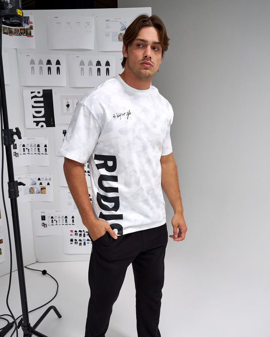 Man wearing a white t-shirt with 'RUDI' printed on it, standing in front of a white wall with graphics.
