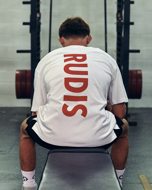 A man sitting on a weight bench in a gym, viewed from behind, wearing a white t-shirt with the brand name 'RUDIS' printed vertically in red on the back.