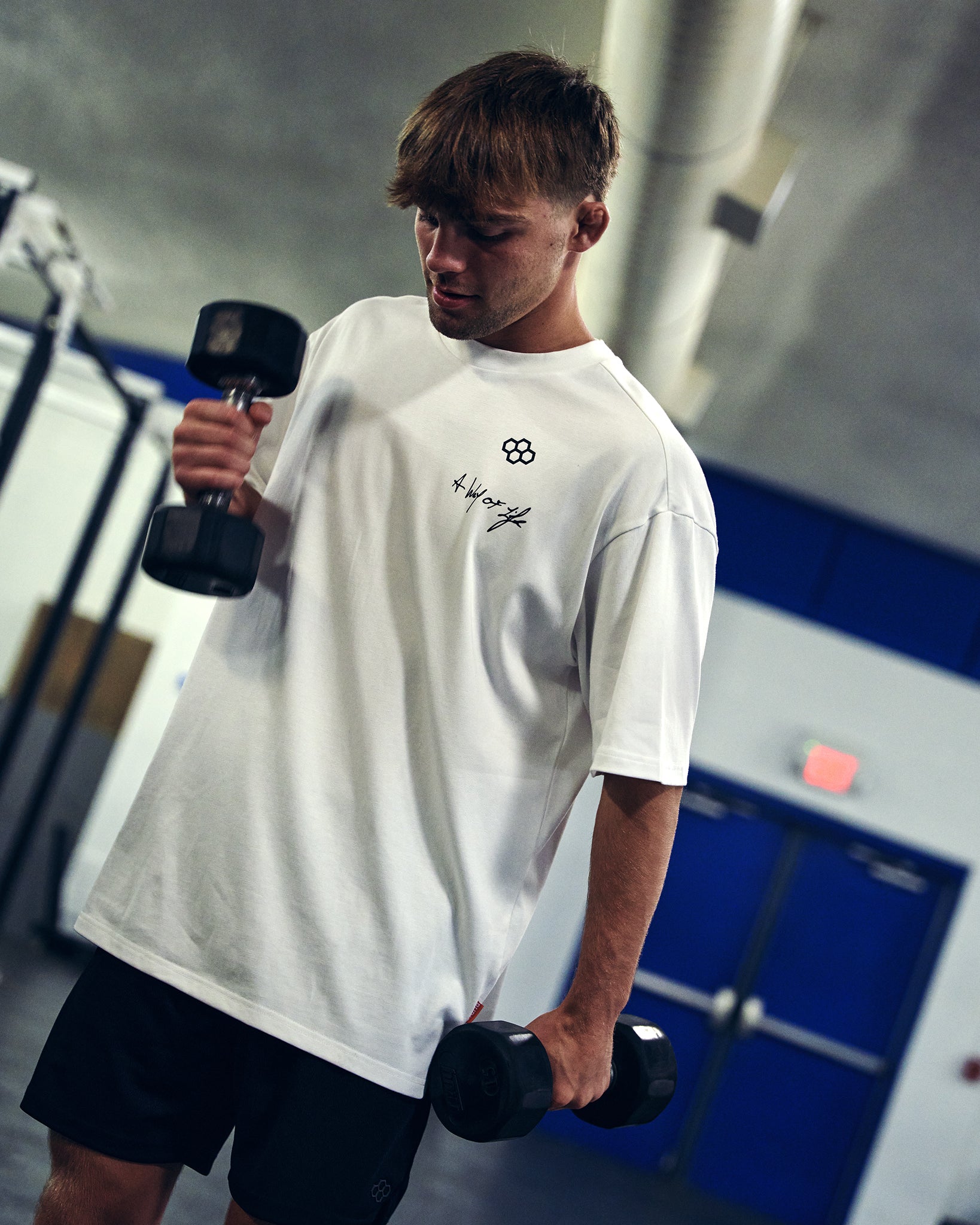 A man in a gym wearing a white crewneck t-shirt with a small black graphic logo on the chest, holding dumbbells.