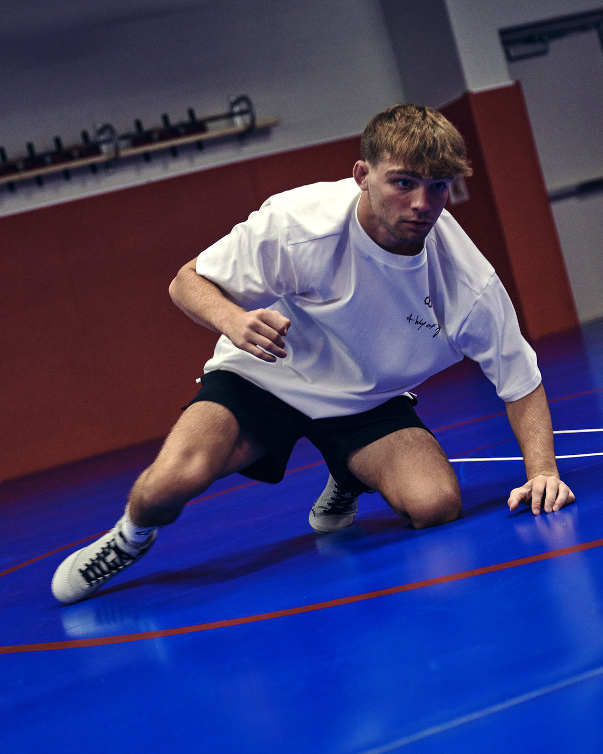 A man in a white t-shirt and black shorts crouches on a blue indoor sports court.
