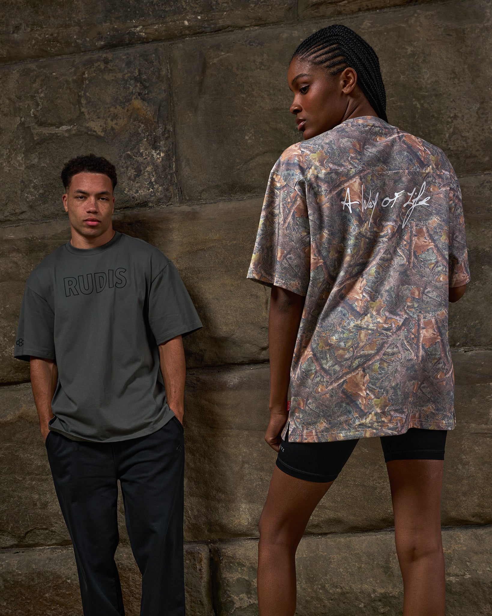 Two people standing against a stone wall, one wearing a gray t-shirt with 'RUPES' branding and the other in a patterned shirt.