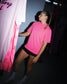 A woman wearing a bright pink short-sleeve t-shirt and black shorts stands in a dimly lit studio or warehouse space, looking to the left where another pink t-shirt is hanging.