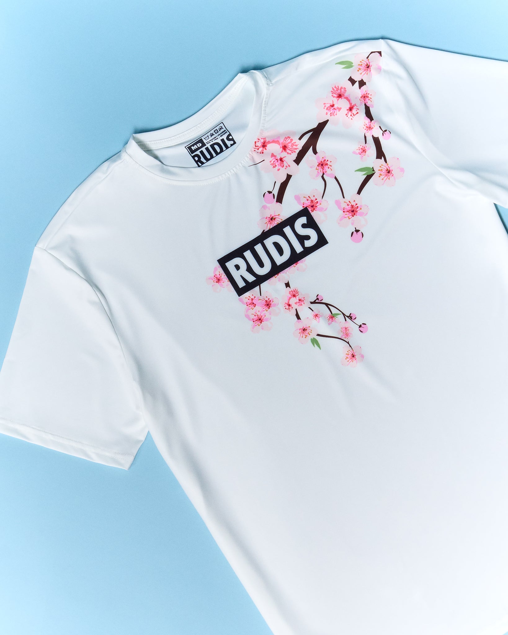 White t-shirt with cherry blossom design and 'RUDIS' logo on a light blue background