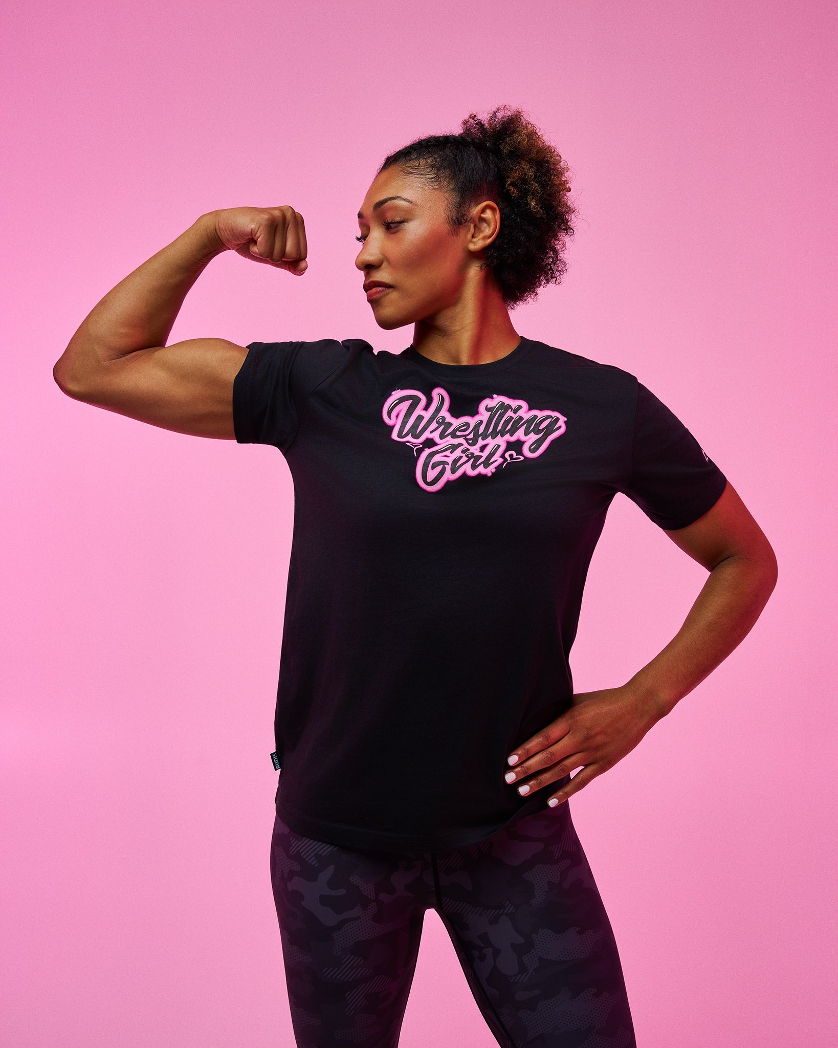 A woman posing in a black short-sleeve t-shirt with 'Wrestling Girl' printed in pink and white script across the chest, paired with dark patterned leggings, set against a solid pink studio background.