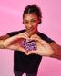 A woman with curly hair wearing a black t-shirt with a pink graphic that reads 'Wrestling Girls', posing with her hands forming a heart shape over the shirt against a solid pink background.