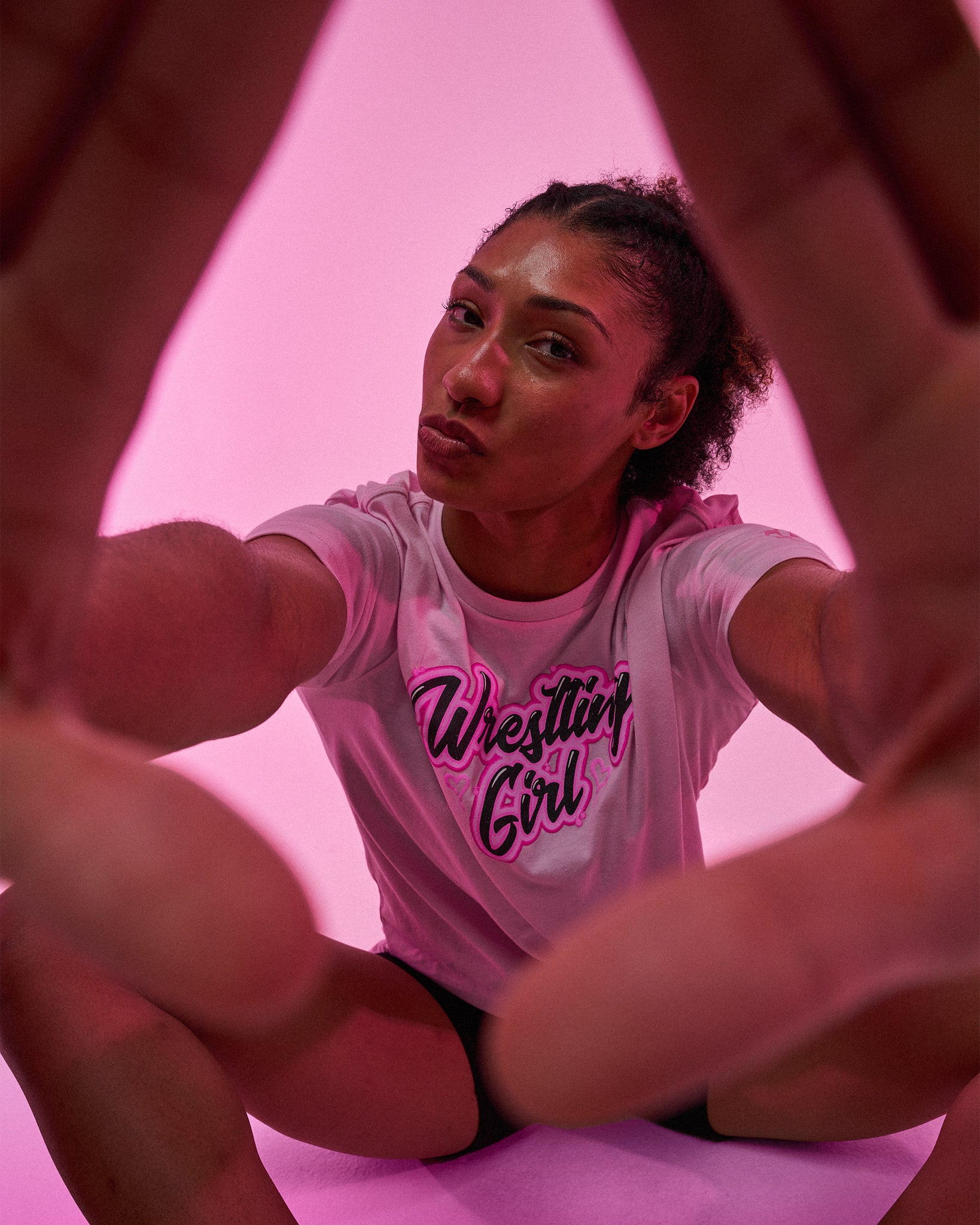 A woman sits cross-legged against a pink background, framed by her hands in the foreground. She wears a light pink graphic T-shirt with the words 'Wrestling Girl' in dark pink and black script.