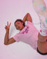 A woman lying on a light pink floor wearing a light pink t-shirt with 'Wrestling Girls' printed in dark pink script, paired with white athletic shoes and black shorts.