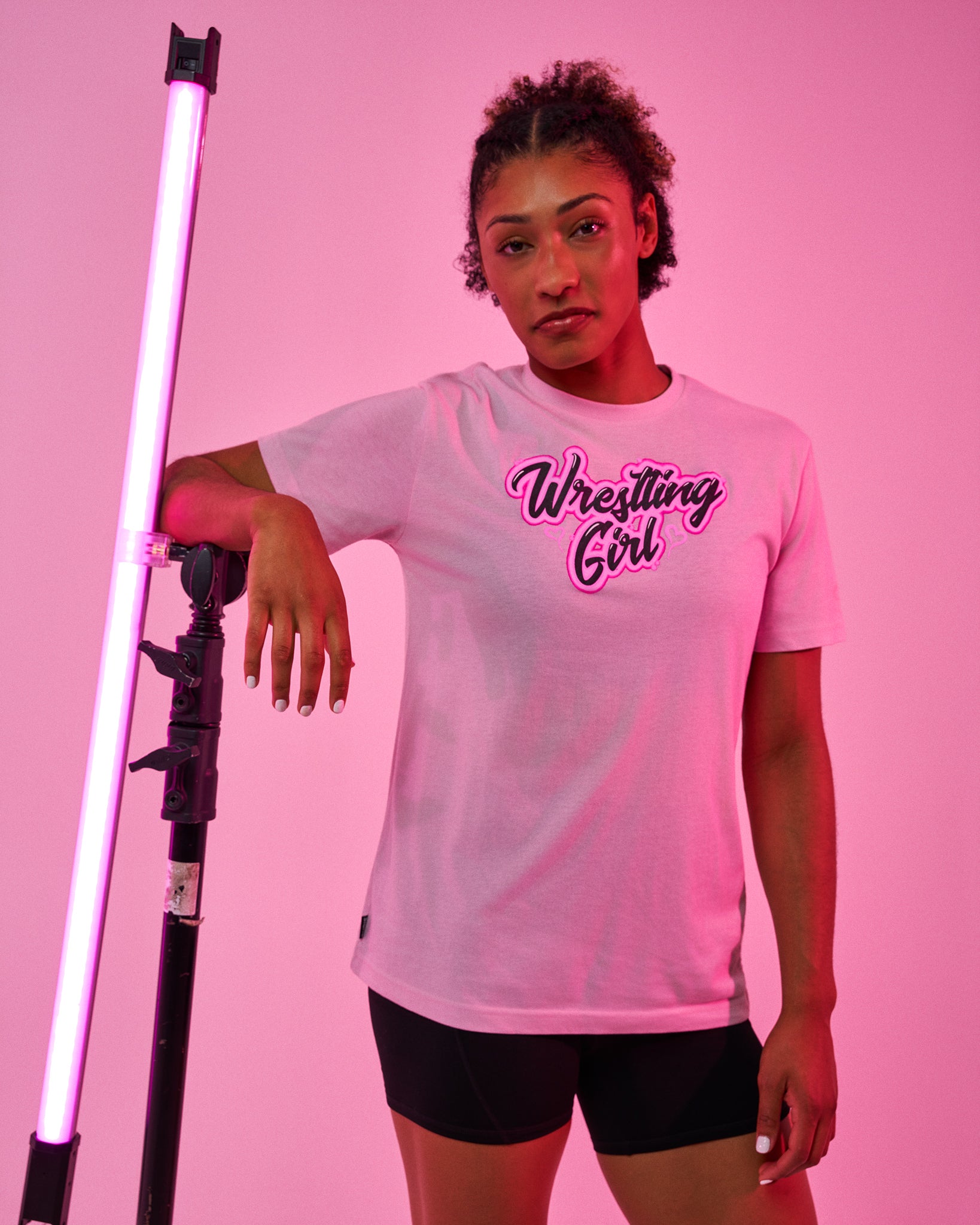 A woman posing in a pink-lit studio wearing a white graphic t-shirt that reads 'Wrestling Girl' in black and pink script, paired with black athletic shorts.