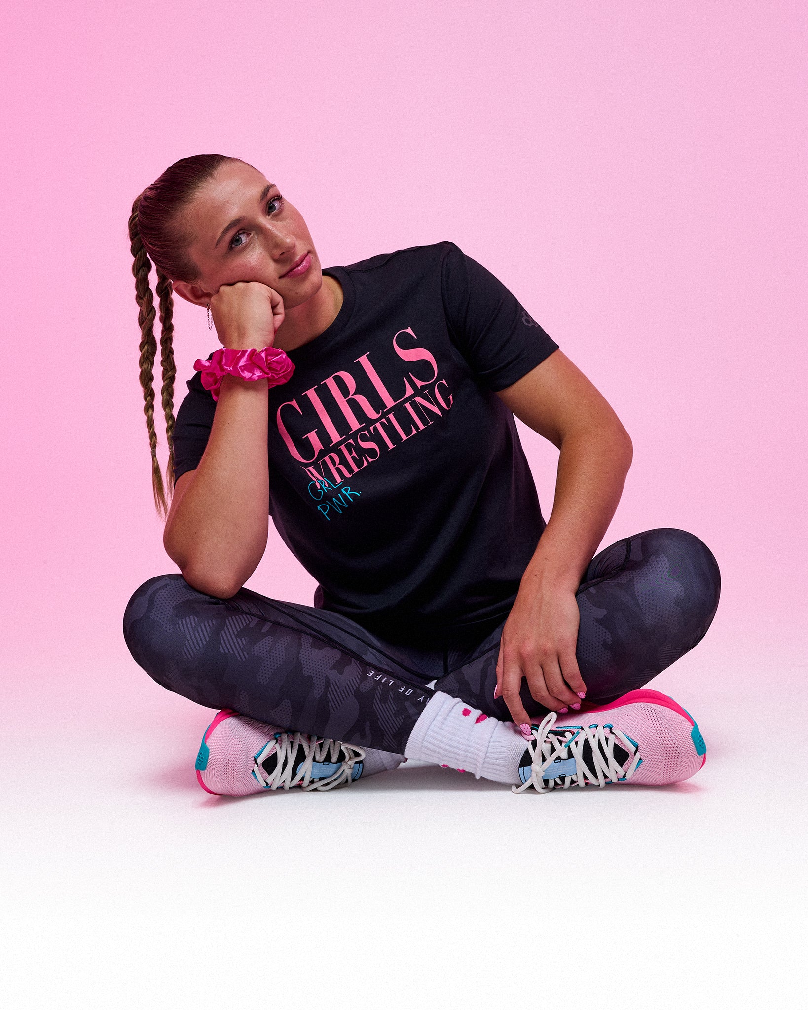 Female athlete seated cross-legged wearing the RUDIS Girl Power Wrestling Elite Super Soft T-Shirt in black, gray camo leggings, and pink wrestling shoes in a pink studio setting.