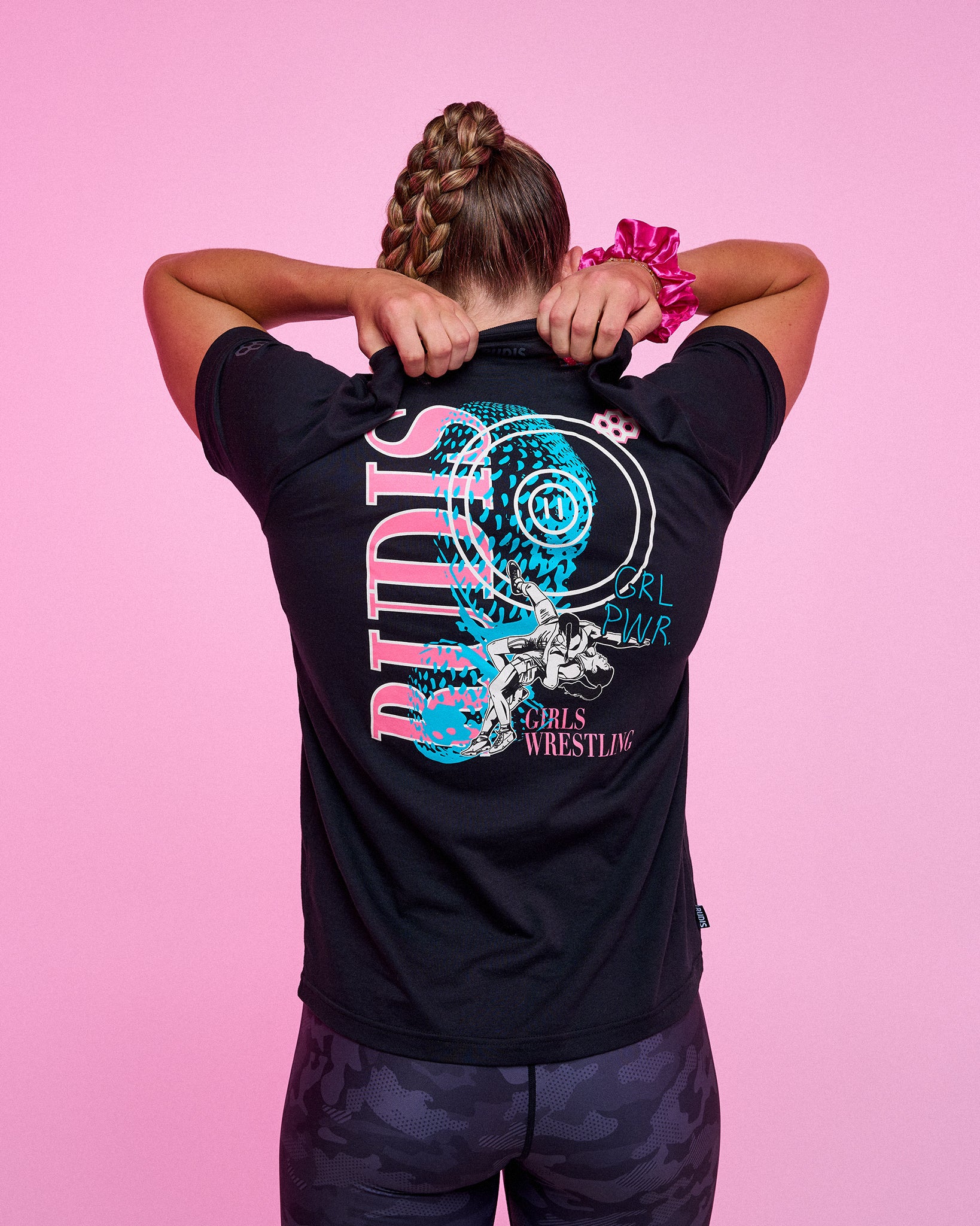 Female athlete wearing the RUDIS Girl Power Wrestling Elite Super Soft T-Shirt in black displays the bold back graphic featuring wrestling artwork against a pink studio background.