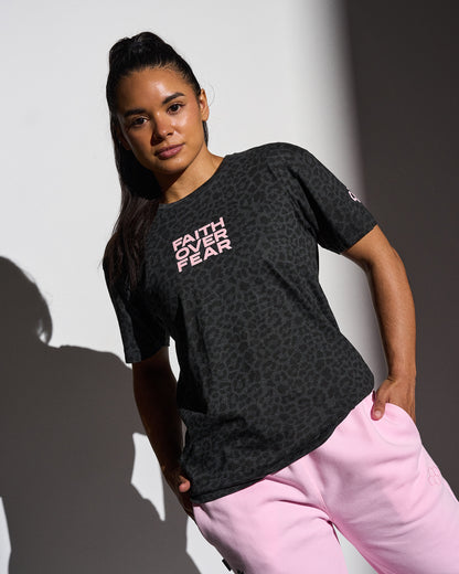 Woman wearing a black animal printed t-shirt with 'Faith Over Fear' text, standing against a white wall.