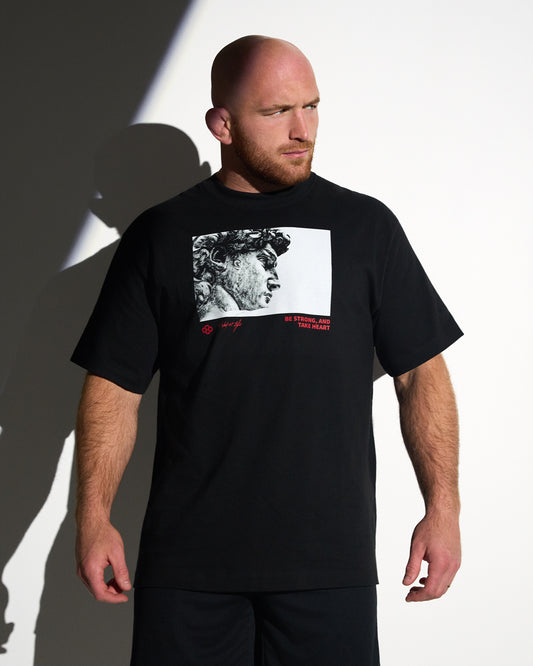 Athlete wearing the RUDIS Be Strong Heavyweight T-Shirt in black featuring a bold classical statue graphic with "Be Strong, and Take Heart" messaging.