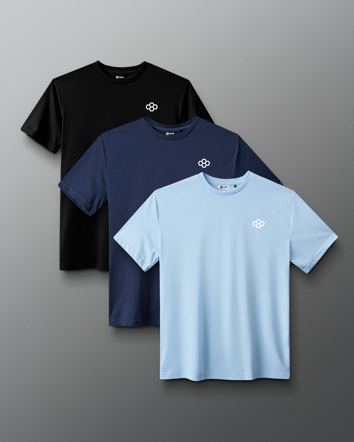 Three short-sleeve crew-neck T-shirts arranged in an overlapping stack on a gray gradient background: black, navy, and light blue, each with a small white geometric chest logo.